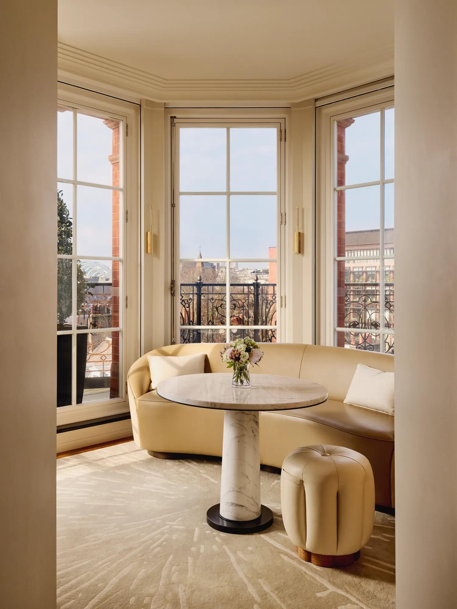 Elegant sitting nook with a curved cream leather sofa and marble-topped round table beside large bay windows, opening onto a balcony with wrought-iron railings and city views.