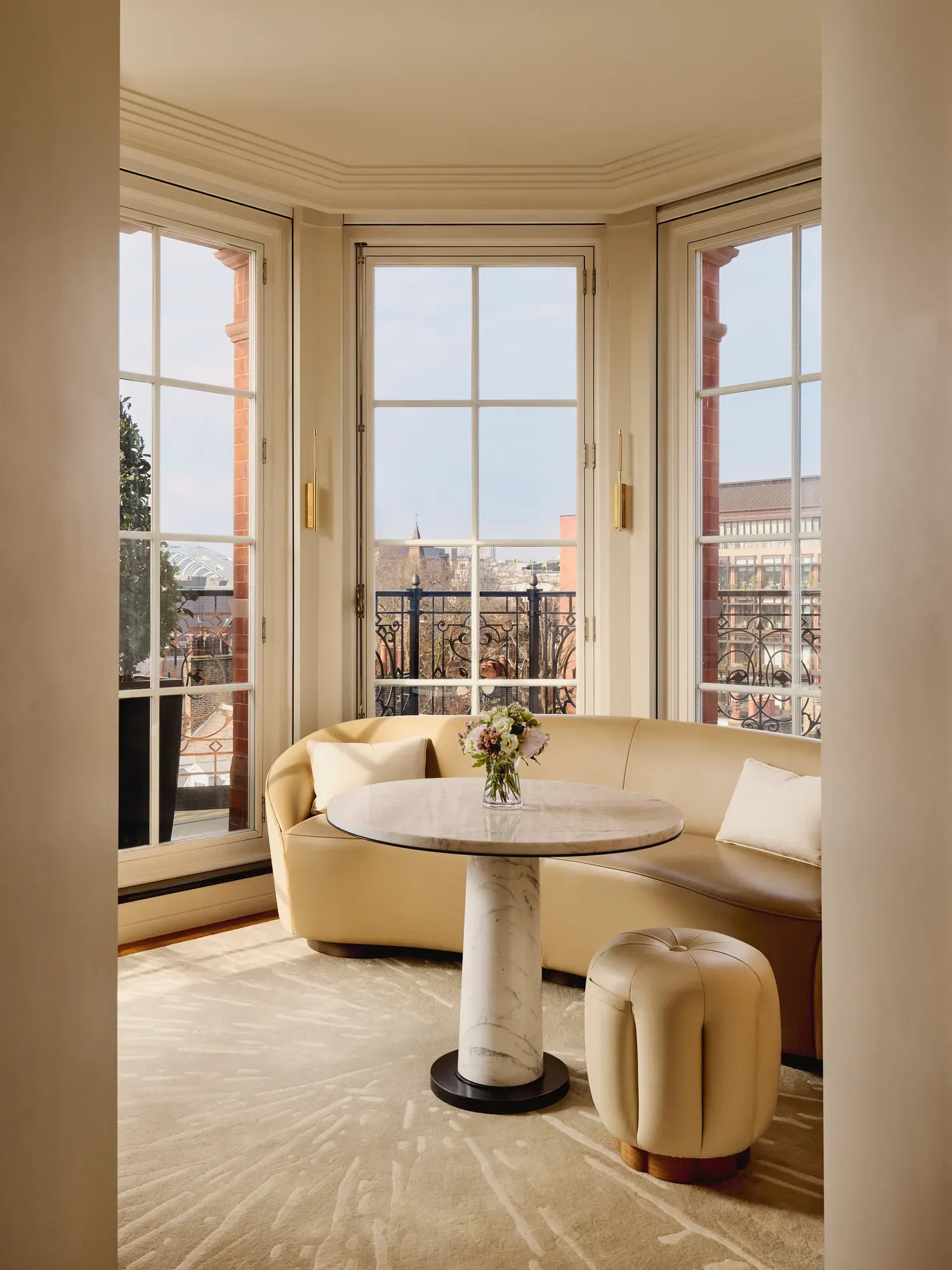 Elegant sitting nook with a curved cream leather sofa and marble-topped round table beside large bay windows, opening onto a balcony with wrought-iron railings and city views.