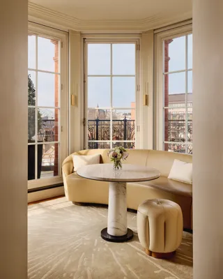 Elegant sitting nook with a curved cream leather sofa and marble-topped round table beside large bay windows, opening onto a balcony with wrought-iron railings and city views.
