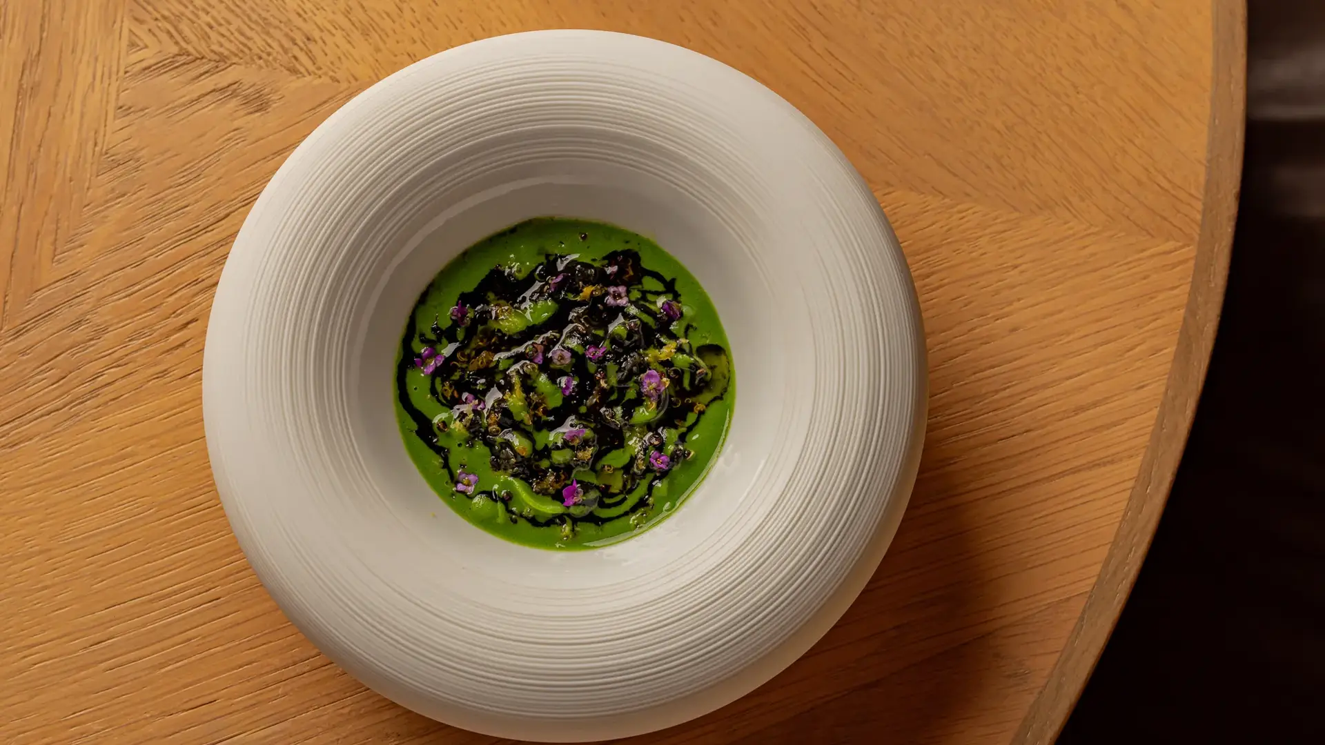 Green soup or sauce topped with herbs, seaweed, and edible flowers, served in a deep white bowl on a wooden table.