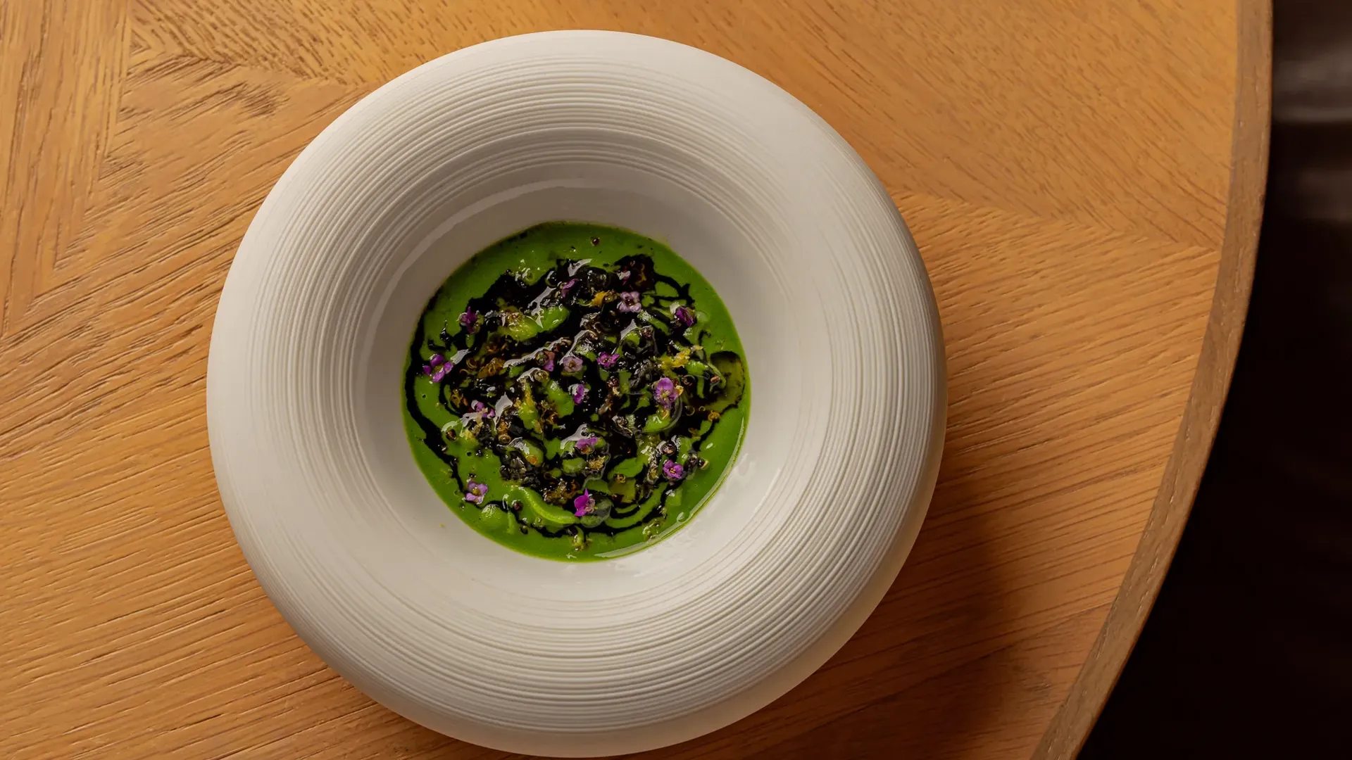 Green soup or sauce topped with herbs, seaweed, and edible flowers, served in a deep white bowl on a wooden table.