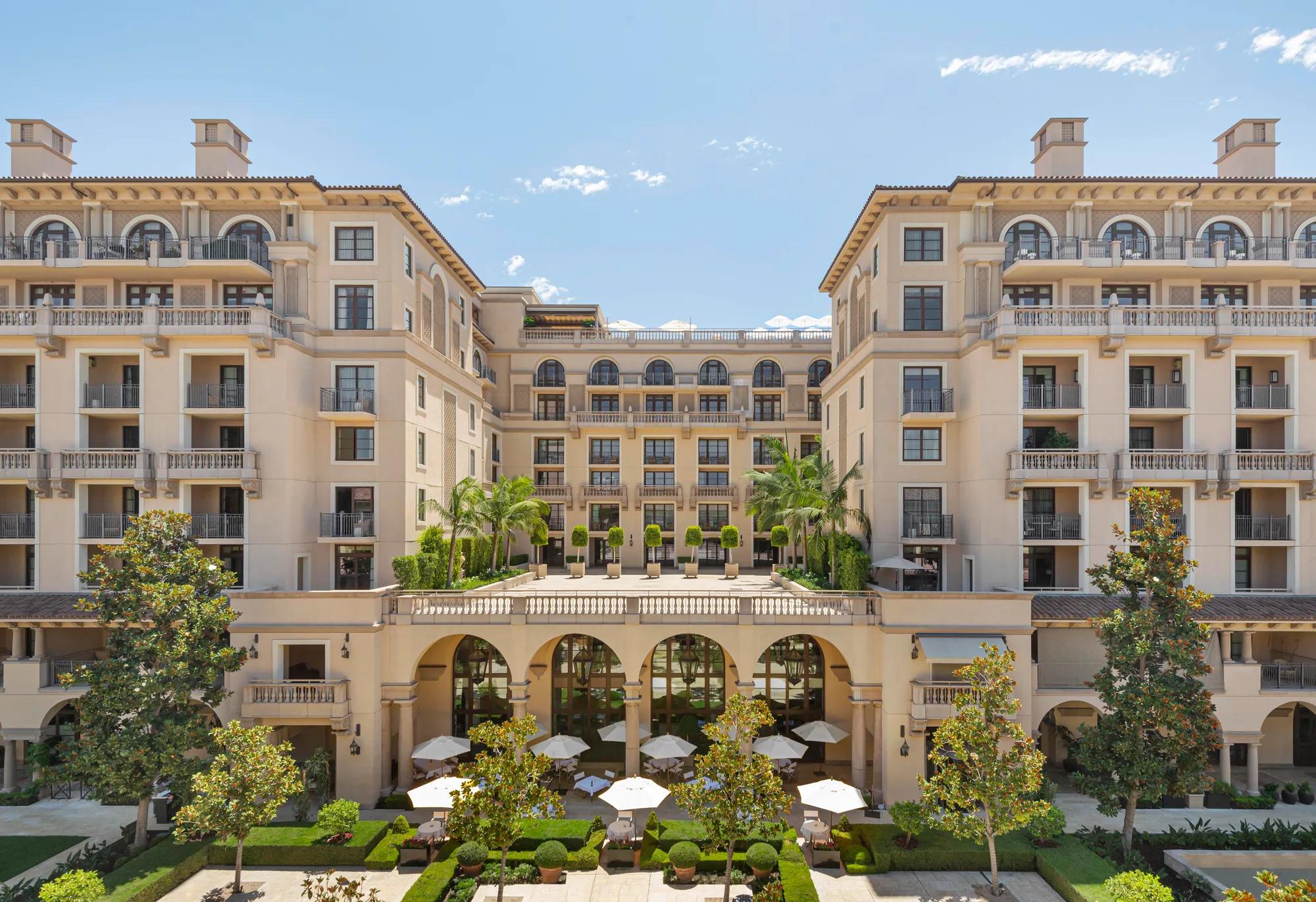 Symmetrical courtyard façade of The Maybourne Beverly Hills featuring grand arches, terrace topiary, and outdoor dining umbrellas.