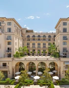 Symmetrical courtyard façade of The Maybourne Beverly Hills featuring grand arches, terrace topiary, and outdoor dining umbrellas.