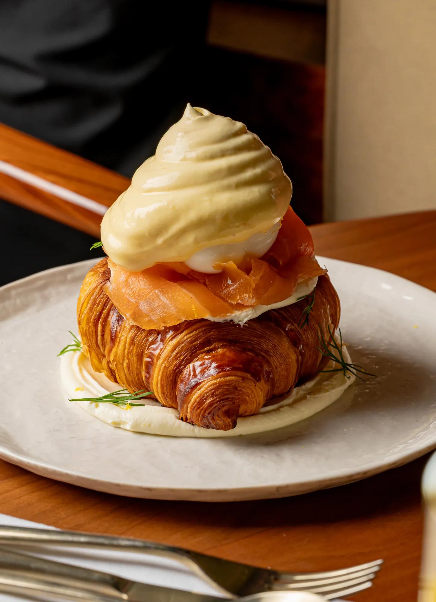 Croissant topped with smoked salmon, poached egg and creamy hollandaise, served on a white plate in warm dining setting