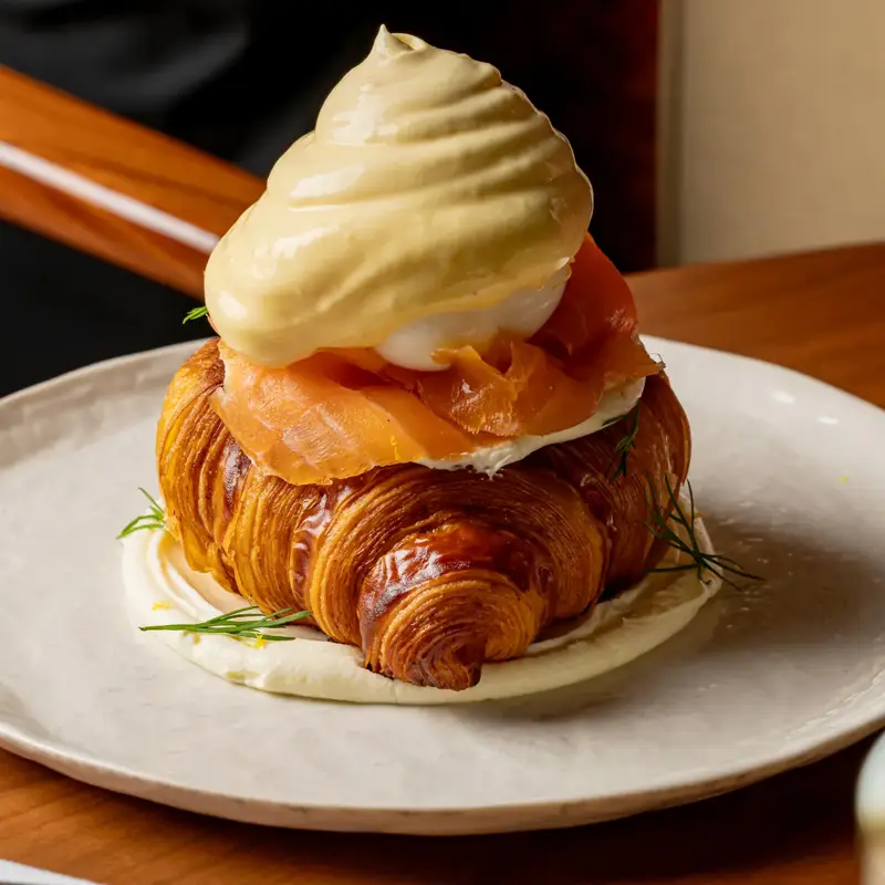 Croissant topped with smoked salmon, poached egg and creamy hollandaise, served on a white plate in warm dining setting