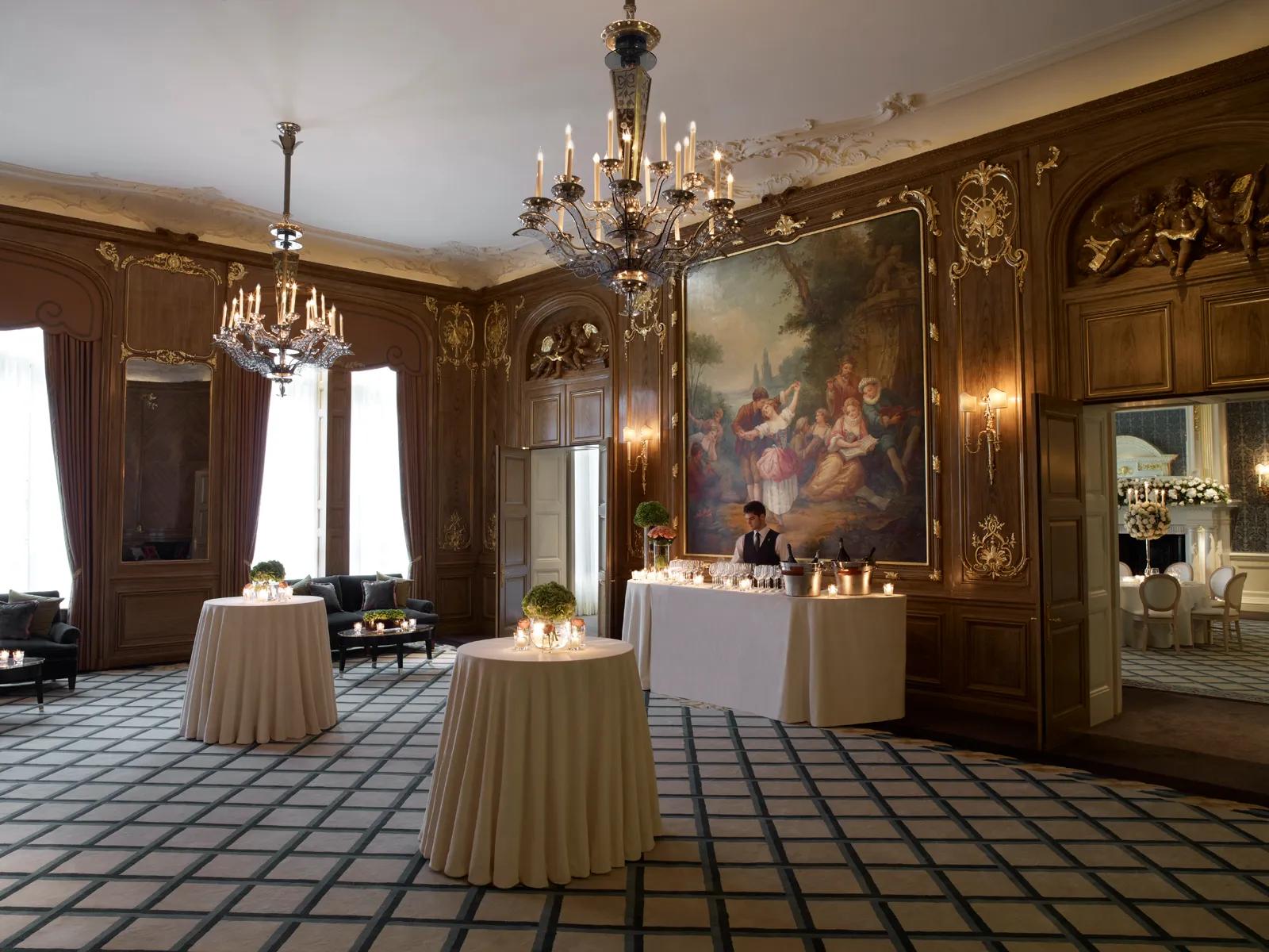 Elegant Claridge’s ballroom set for a daytime reception with round cocktail tables draped in white linen, candles and floral arrangements, and a bartender at a drinks table beneath a large classical painting.
