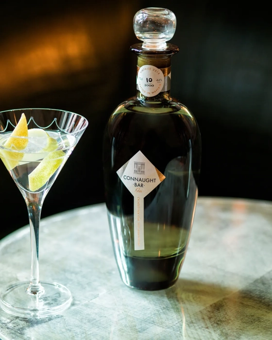 Martini glass with lemon twists beside a dark Connaught Bar gin bottle on a reflective table.