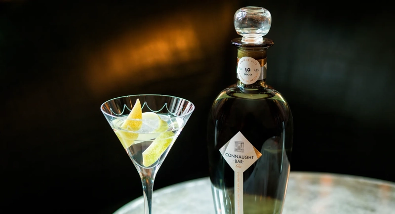 Martini glass with lemon twists beside a dark Connaught Bar gin bottle on a reflective table.