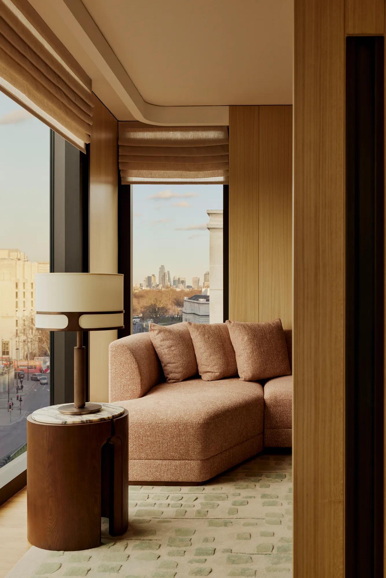 Floor-to-ceiling window seating with a curved upholstered sofa, side table and lamp, overlooking a city skyline and treetops.