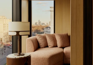 Floor-to-ceiling window seating with a curved upholstered sofa, side table and lamp, overlooking a city skyline and treetops.