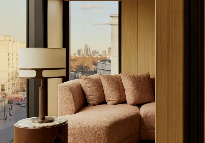 Floor-to-ceiling window seating with a curved upholstered sofa, side table and lamp, overlooking a city skyline and treetops.
