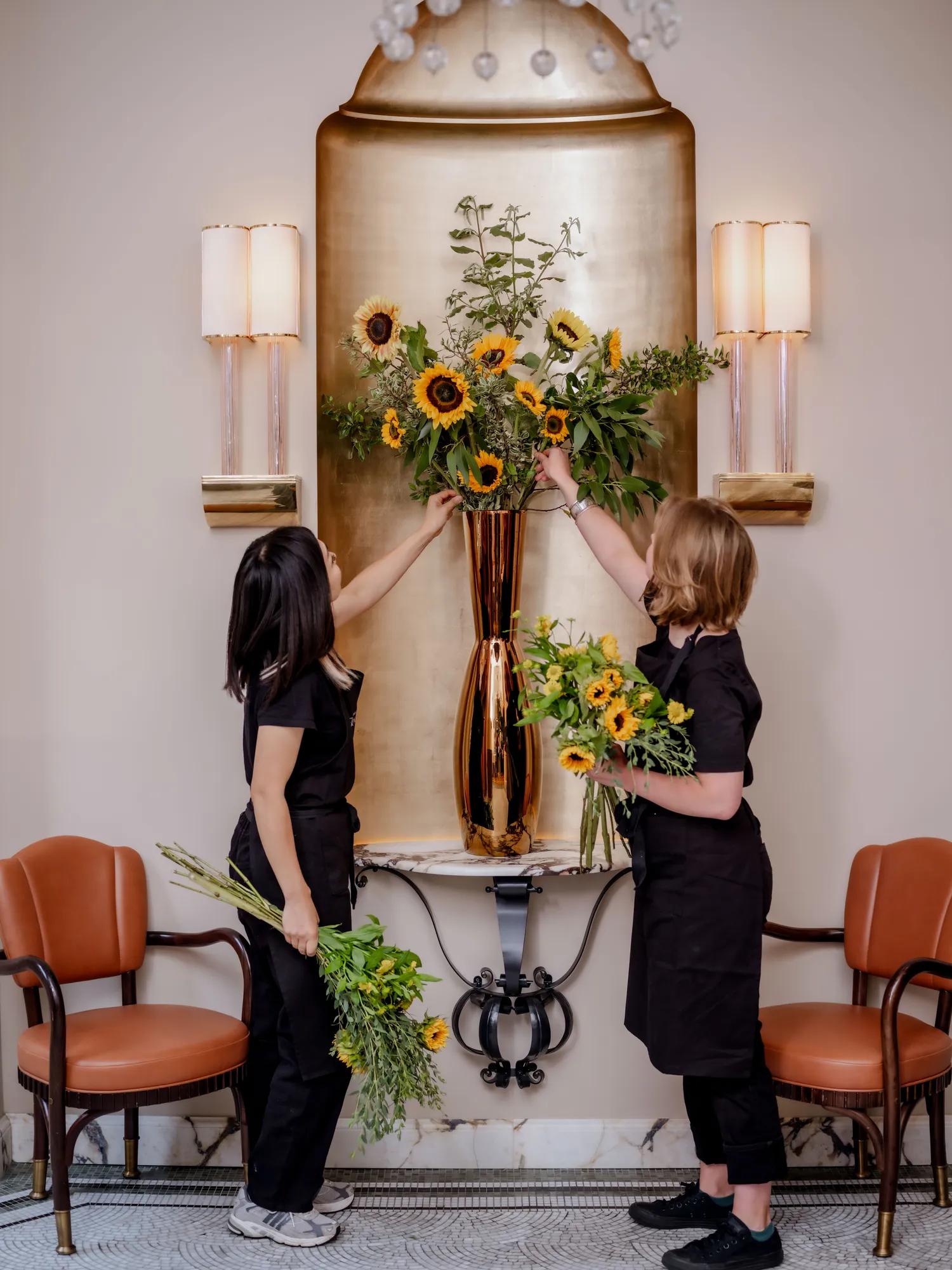Two florists arrange sunflowers and greenery in a tall gold vase on a wall-mounted console between leather chairs.