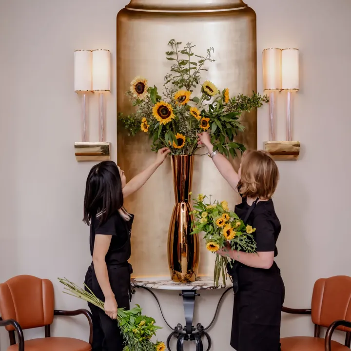 Two florists arrange sunflowers and greenery in a tall gold vase on a wall-mounted console between leather chairs.