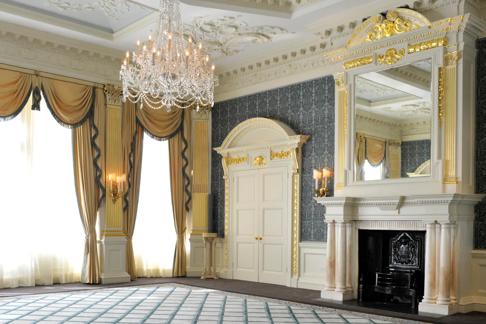 Grand ballroom with ornate gold detailing, crystal chandelier, tall arched windows draped in gold curtains, and an elegant marble fireplace beneath a large gilded mirror.