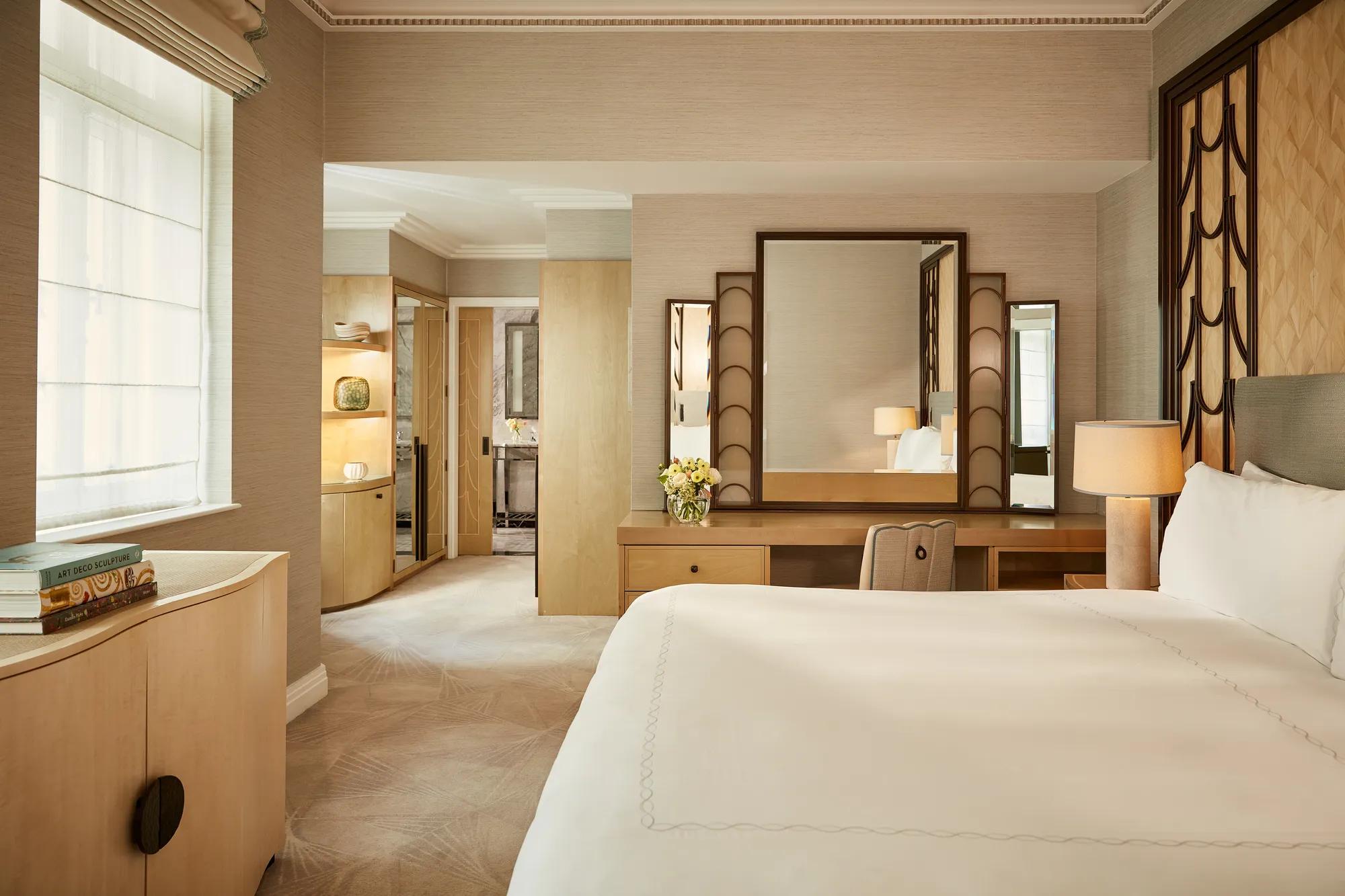 Serene bedroom with a king bed, geometric Art Deco headboard, and a wooden dressing table with a large mirror, softly lit by natural light and warm lamps, leading to a marble bathroom through an open doorway.