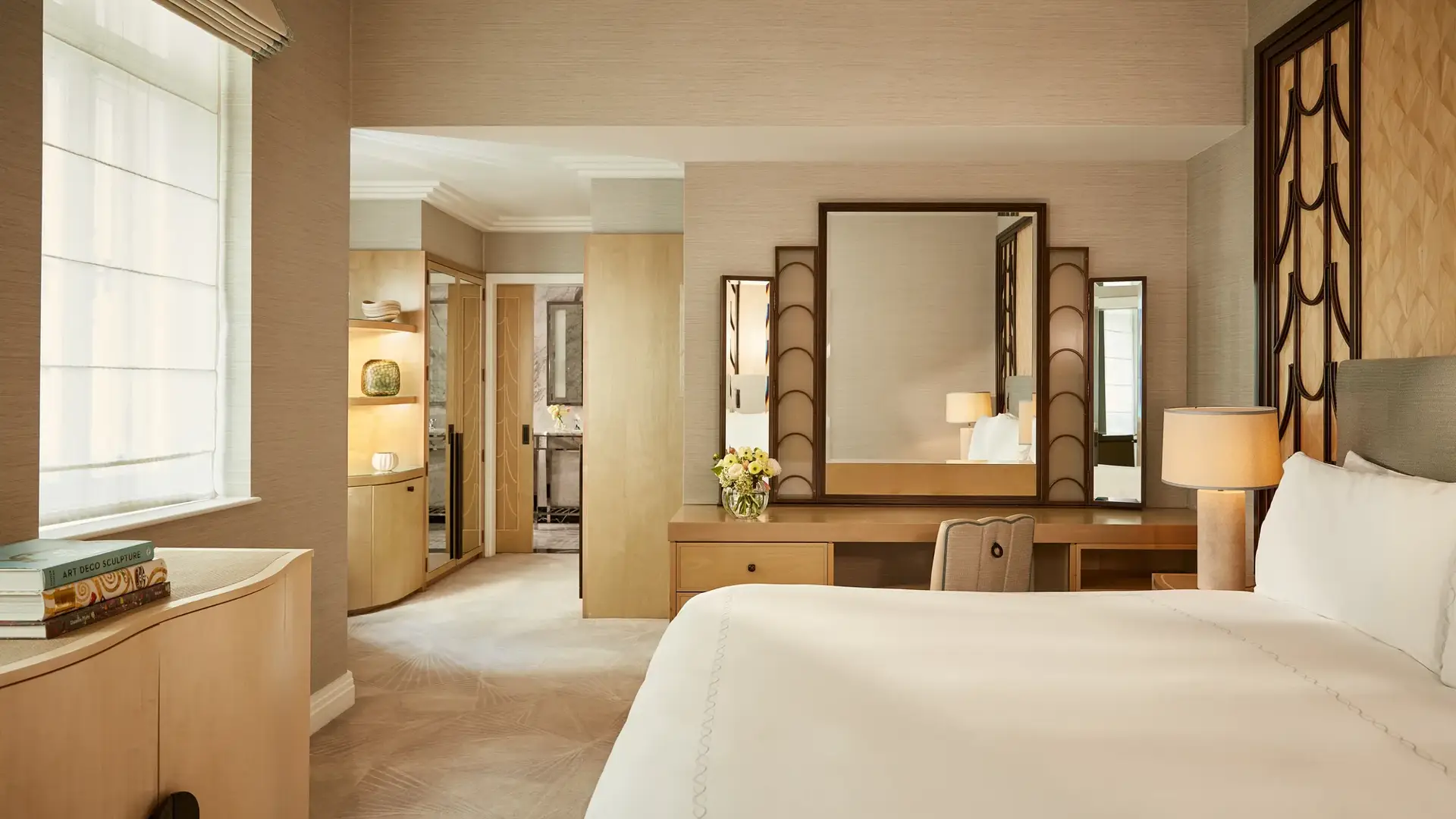 Serene bedroom with a king bed, geometric Art Deco headboard, and a wooden dressing table with a large mirror, softly lit by natural light and warm lamps, leading to a marble bathroom through an open doorway.