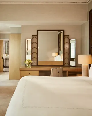 Serene bedroom with a king bed, geometric Art Deco headboard, and a wooden dressing table with a large mirror, softly lit by natural light and warm lamps, leading to a marble bathroom through an open doorway.