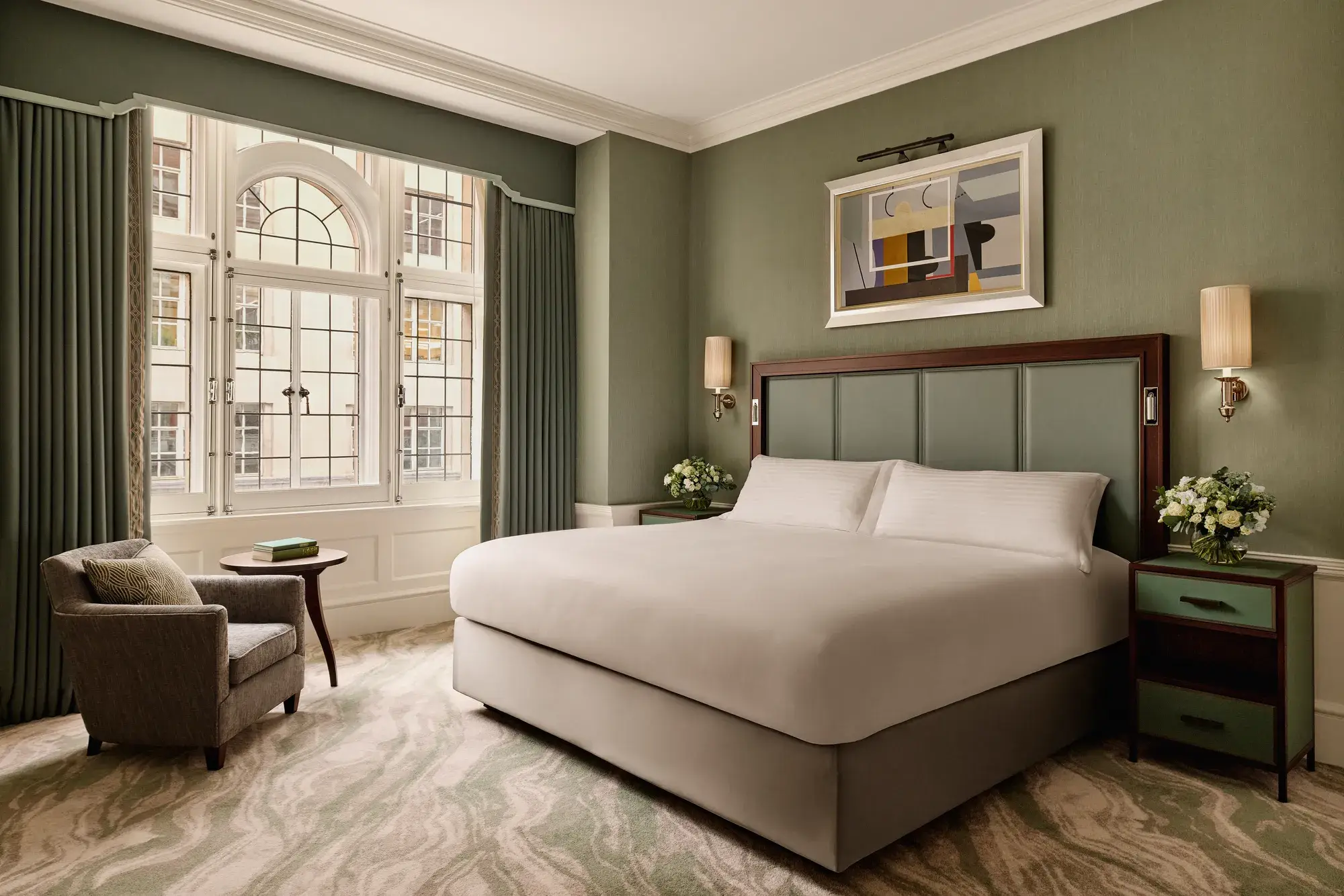 Elegant bedroom with king bed, green walls, armchair, and large arched window.