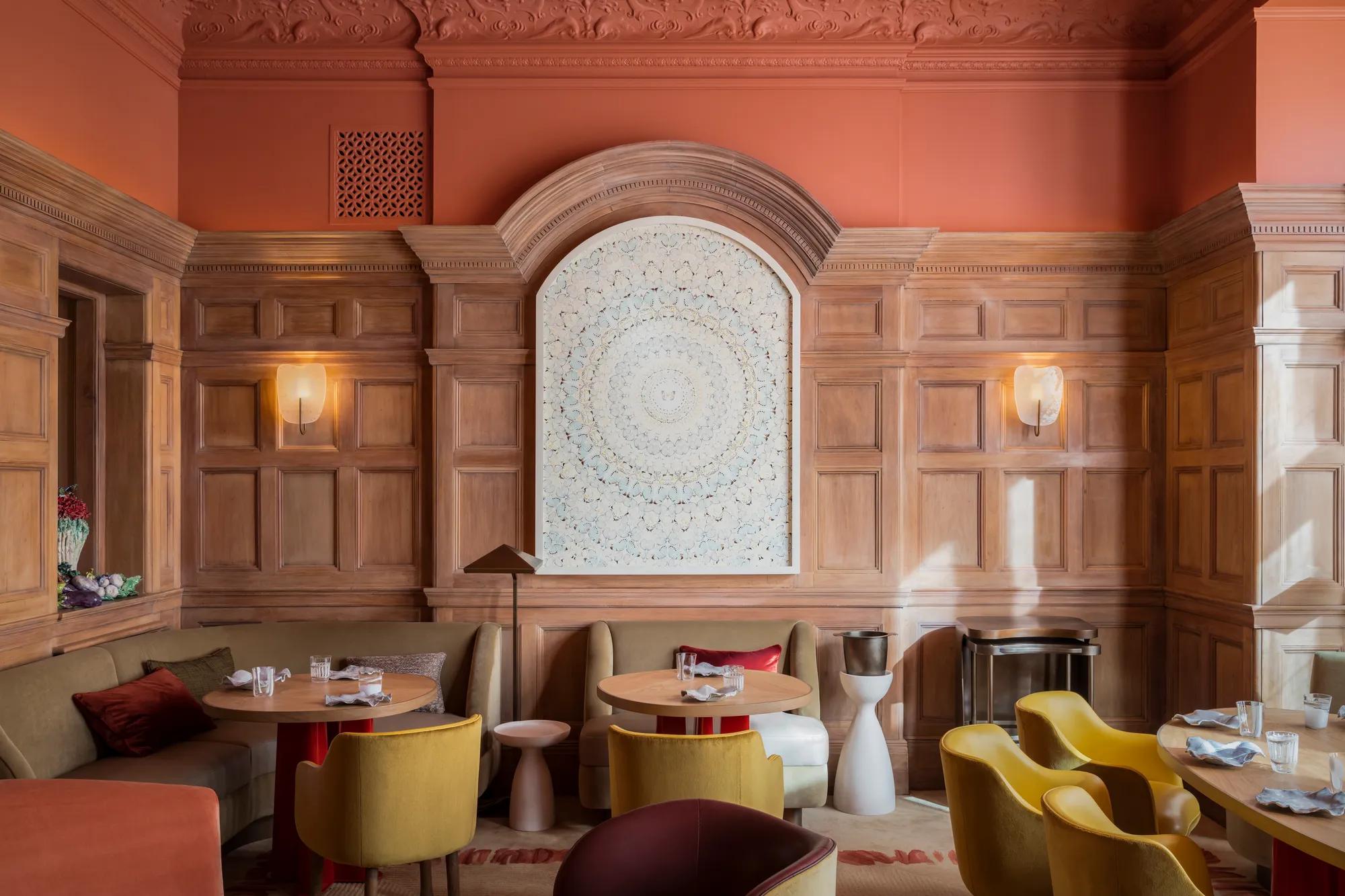 Elegant dining room with wood panelled walls, round tables, mustard chairs, and decorative arched wall feature.
