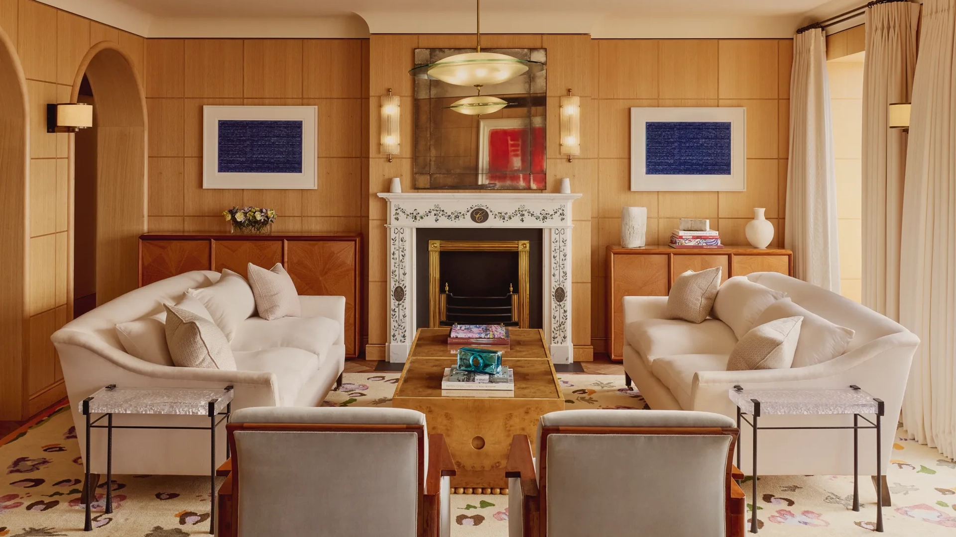 Elegant living room with two cream sofas facing each other, a marble fireplace with decorative tiles, and wood-paneled walls featuring framed abstract art in blue.