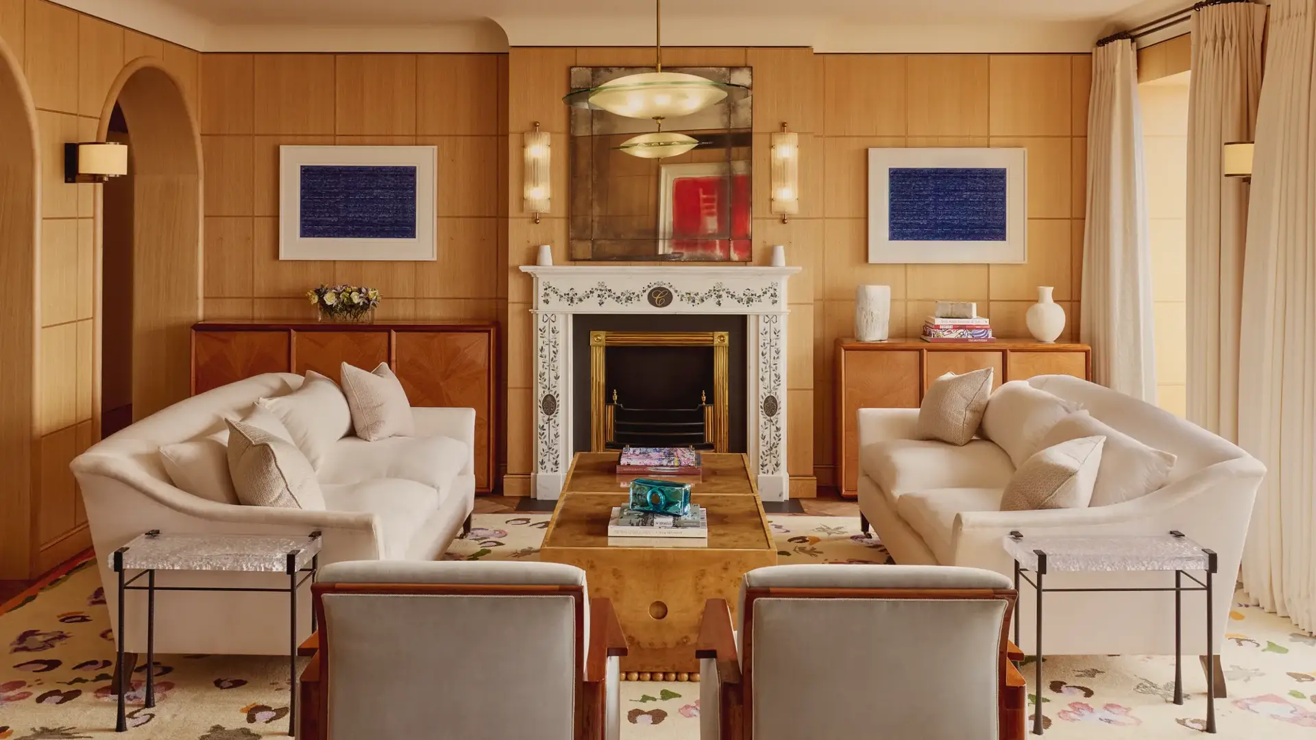 Elegant living room with two cream sofas facing each other, a marble fireplace with decorative tiles, and wood-paneled walls featuring framed abstract art in blue.