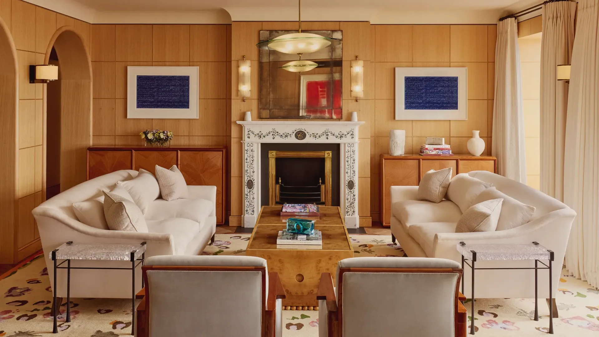 Elegant living room with two cream sofas facing each other, a marble fireplace with decorative tiles, and wood-paneled walls featuring framed abstract art in blue.