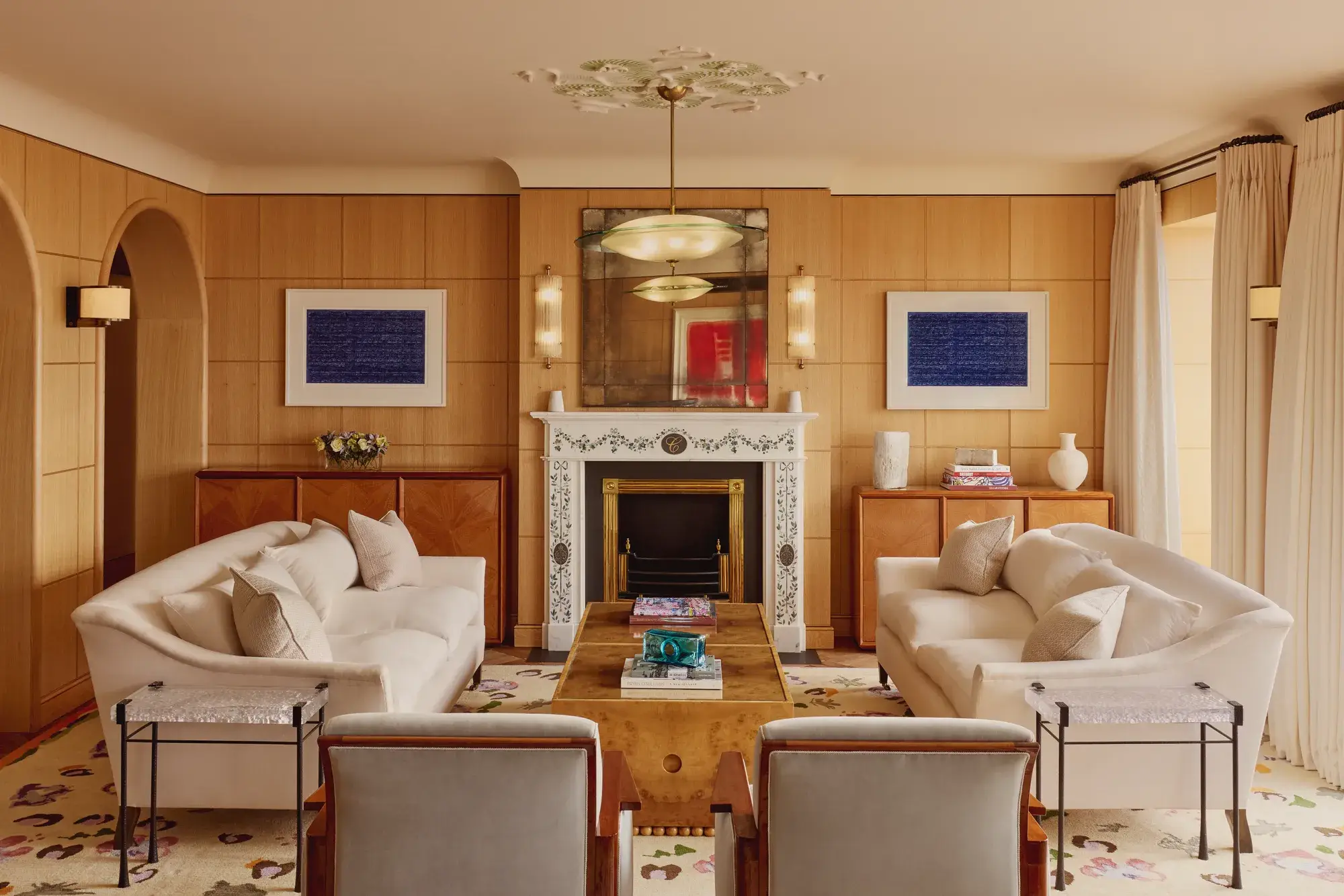 Elegant living room with two cream sofas facing each other, a marble fireplace with decorative tiles, and wood-paneled walls featuring framed abstract art in blue.