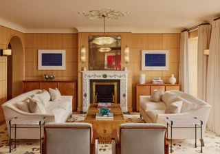 Elegant living room with two cream sofas facing each other, a marble fireplace with decorative tiles, and wood-paneled walls featuring framed abstract art in blue.