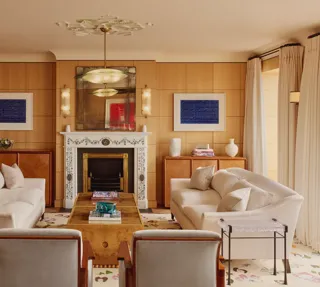 Elegant living room with two cream sofas facing each other, a marble fireplace with decorative tiles, and wood-paneled walls featuring framed abstract art in blue.