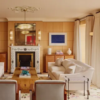 Elegant living room with two cream sofas facing each other, a marble fireplace with decorative tiles, and wood-paneled walls featuring framed abstract art in blue.