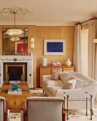 Elegant living room with two cream sofas facing each other, a marble fireplace with decorative tiles, and wood-paneled walls featuring framed abstract art in blue.