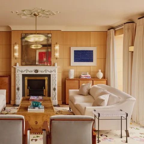 Elegant living room with two cream sofas facing each other, a marble fireplace with decorative tiles, and wood-paneled walls featuring framed abstract art in blue.