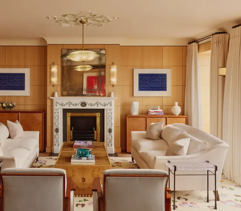 Elegant living room with two cream sofas facing each other, a marble fireplace with decorative tiles, and wood-paneled walls featuring framed abstract art in blue.