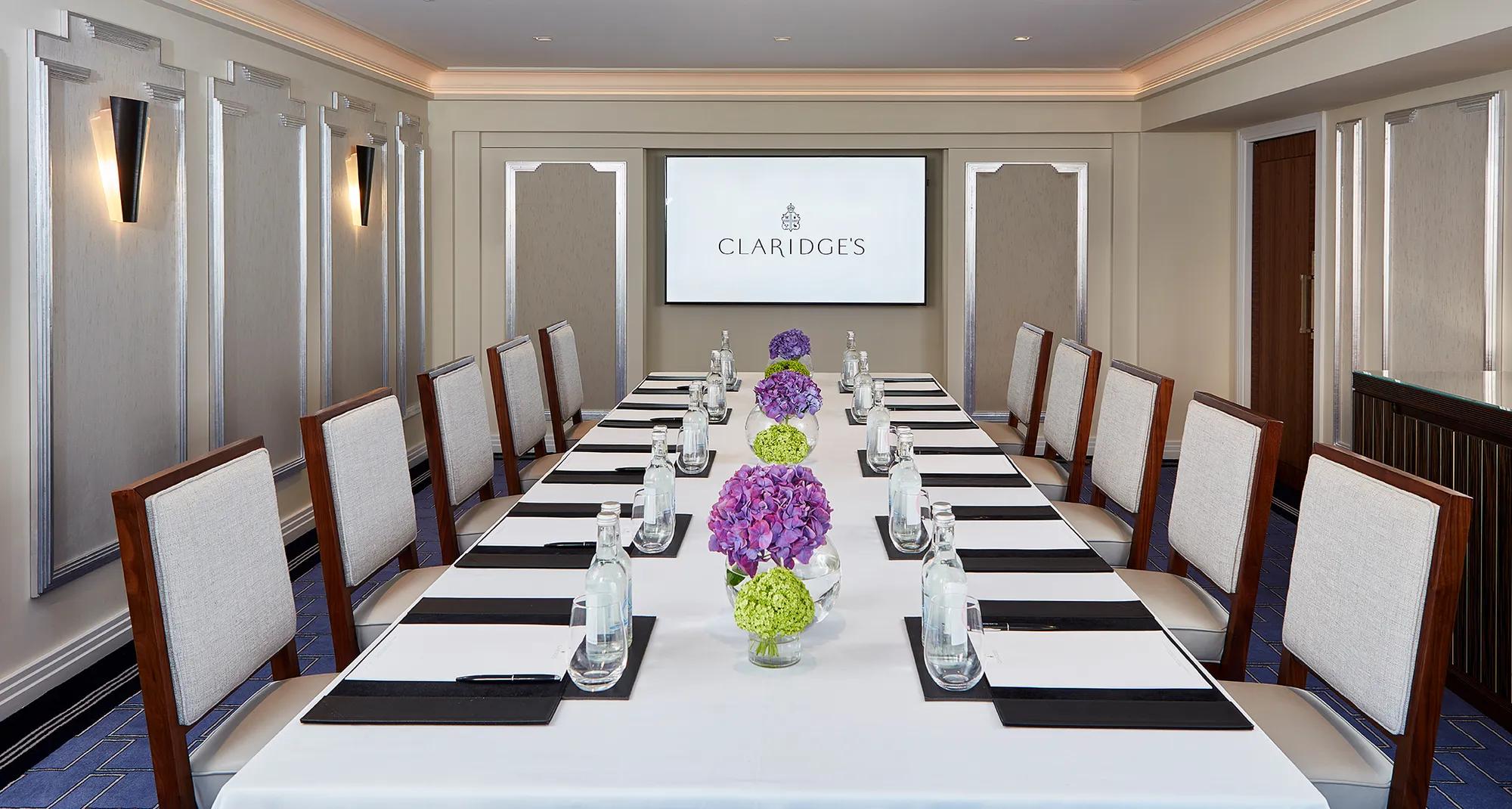 A modern meeting room at Claridge’s featuring a long rectangular table set with water bottles, notepads, and vases of purple and green hydrangeas, with upholstered chairs and a presentation screen at the far end.