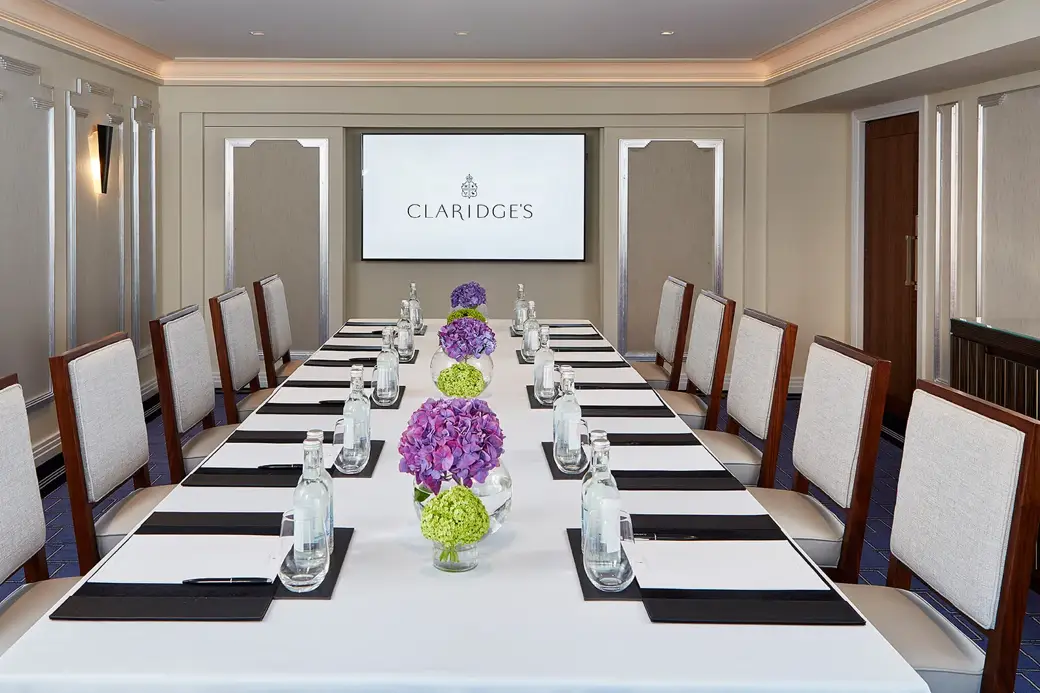 A modern meeting room at Claridge’s featuring a long rectangular table set with water bottles, notepads, and vases of purple and green hydrangeas, with upholstered chairs and a presentation screen at the far end.