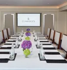 A modern meeting room at Claridge’s featuring a long rectangular table set with water bottles, notepads, and vases of purple and green hydrangeas, with upholstered chairs and a presentation screen at the far end.