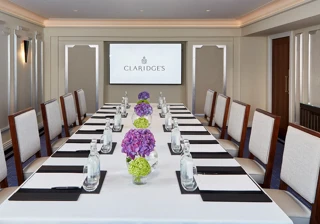 A modern meeting room at Claridge’s featuring a long rectangular table set with water bottles, notepads, and vases of purple and green hydrangeas, with upholstered chairs and a presentation screen at the far end.