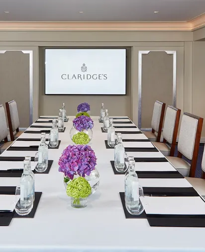 A modern meeting room at Claridge’s featuring a long rectangular table set with water bottles, notepads, and vases of purple and green hydrangeas, with upholstered chairs and a presentation screen at the far end.