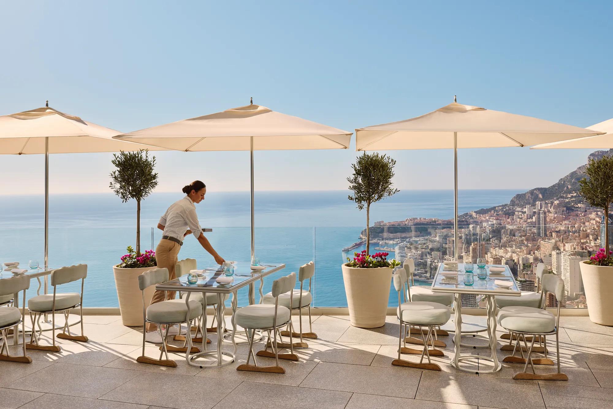 Serveur préparant une table sur une terrasse avec parasols, face à la mer et à une ville côtière.