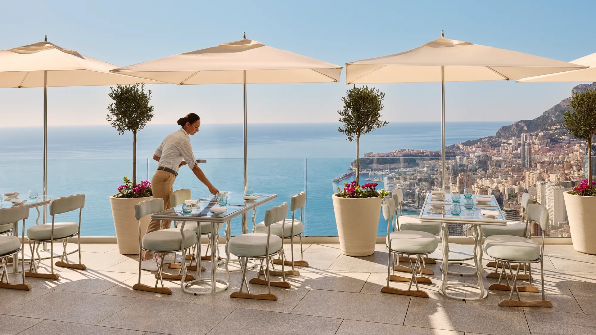 Restaurant terrace with server setting a table beneath umbrellas, overlooking the sea and distant coastal city.