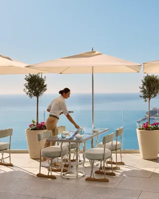 Serveur préparant une table sur une terrasse avec parasols, face à la mer et à une ville côtière.