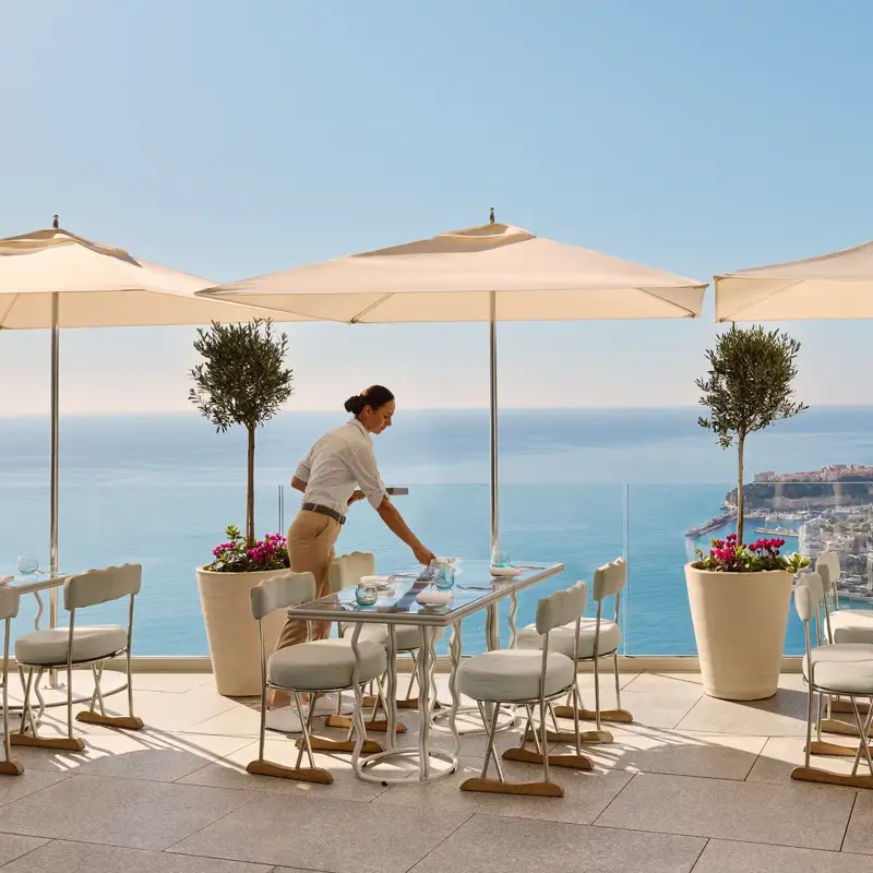Restaurant terrace with server setting a table beneath umbrellas, overlooking the sea and distant coastal city.