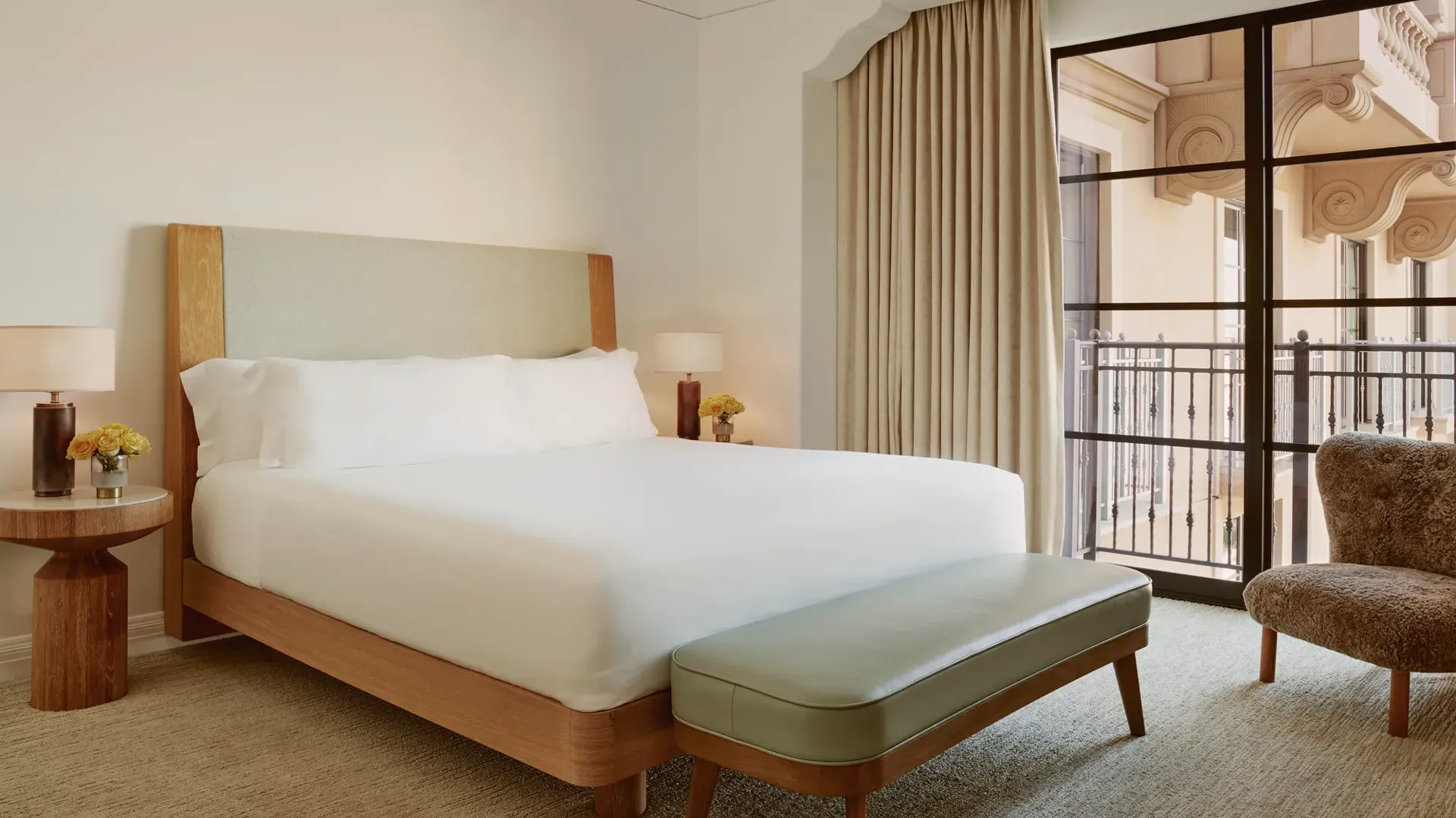 Minimal bedroom with upholstered bed, sage bench, shearling chair, and floor‑to‑ceiling window to balcony.
