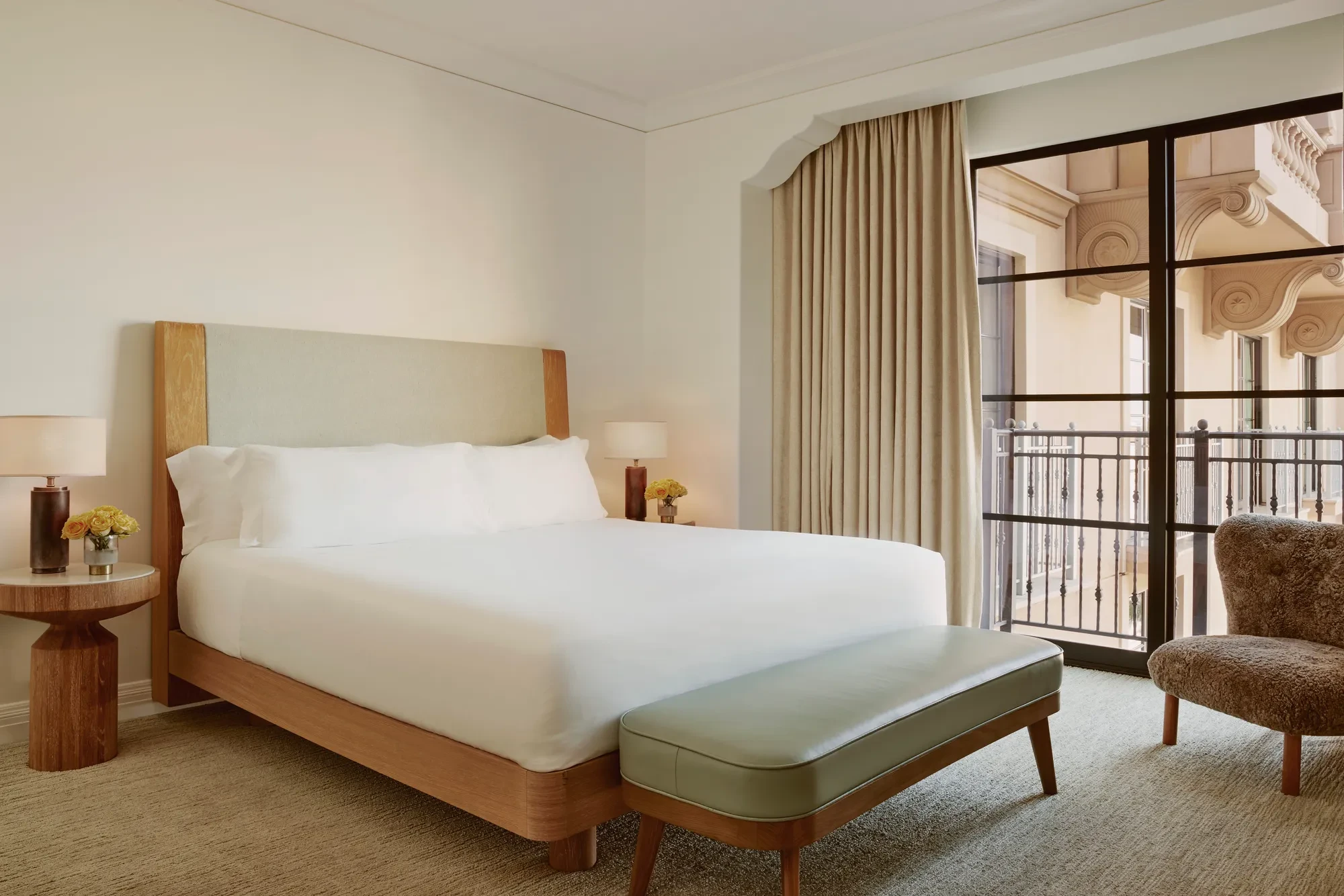 Minimal bedroom with upholstered bed, sage bench, shearling chair, and floor‑to‑ceiling window to balcony.