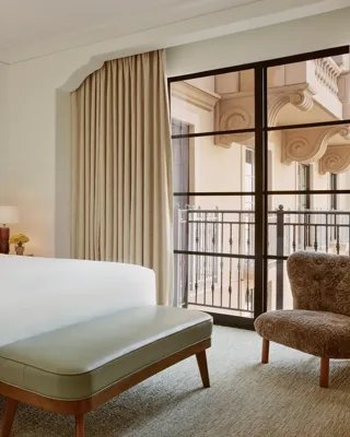 Minimal bedroom with upholstered bed, sage bench, shearling chair, and floor‑to‑ceiling window to balcony.