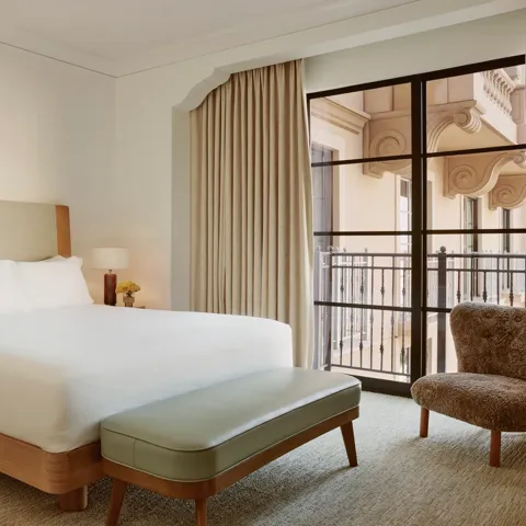Minimal bedroom with upholstered bed, sage bench, shearling chair, and floor‑to‑ceiling window to balcony.