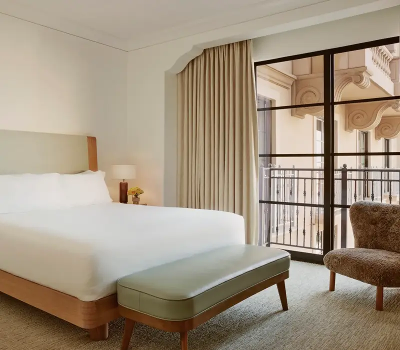 Minimal bedroom with upholstered bed, sage bench, shearling chair, and floor‑to‑ceiling window to balcony.