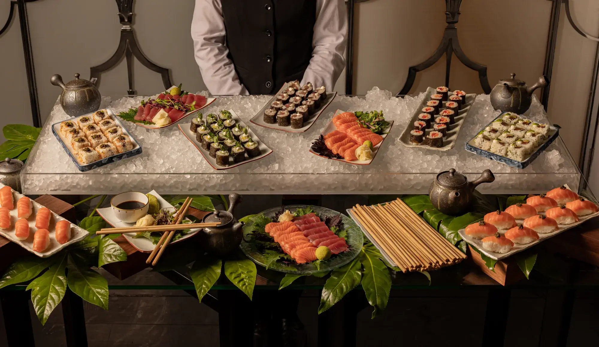 A server stands behind an elegant sushi display featuring an assortment of nigiri, sashimi, and sushi rolls arranged on ice and leafy garnishes, with teapots, soy sauce, and chopsticks neatly placed on the table.