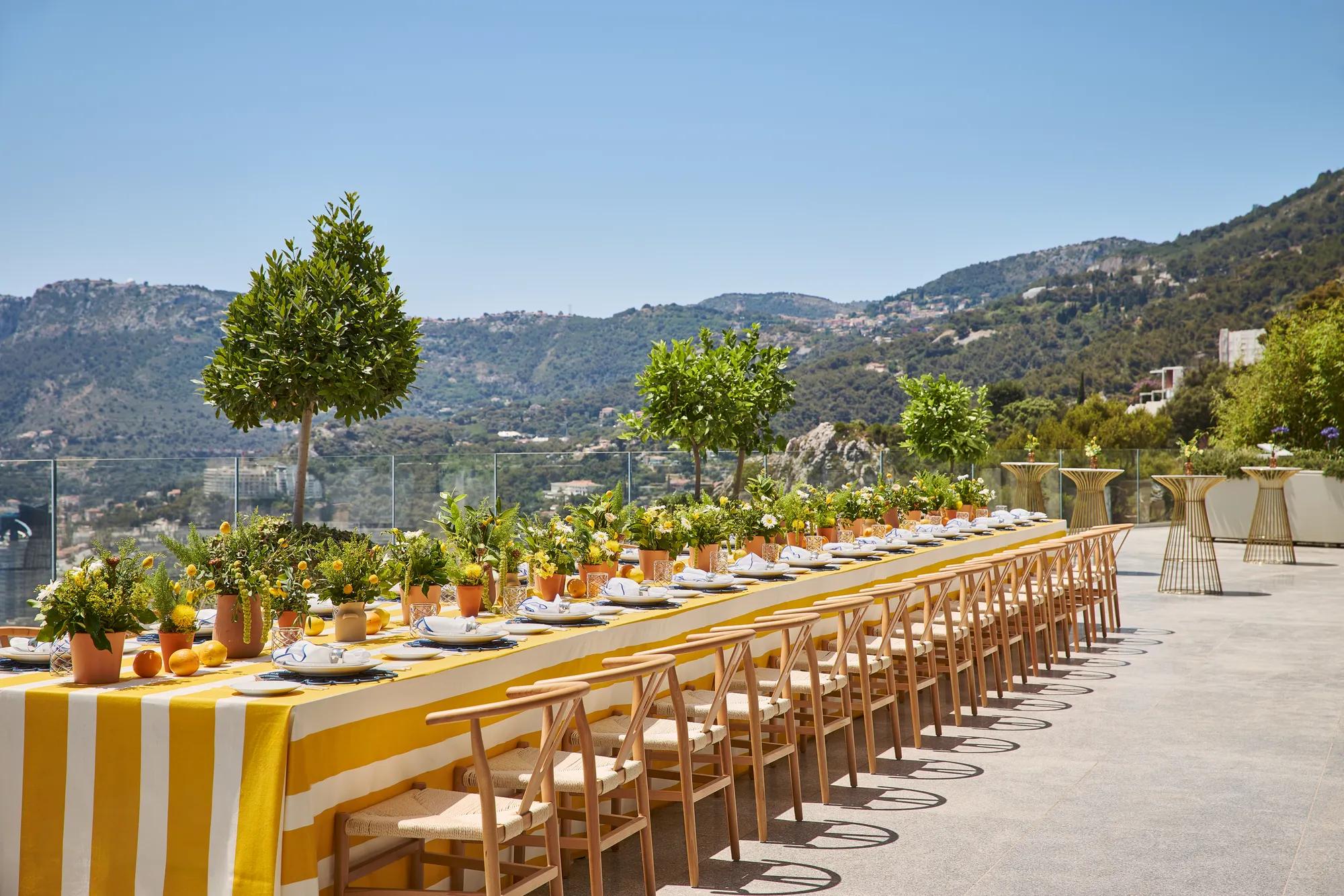 Longue table dressée en extérieur avec nappes jaunes, chaises en bois, plantes en pots et vue sur les montagnes.