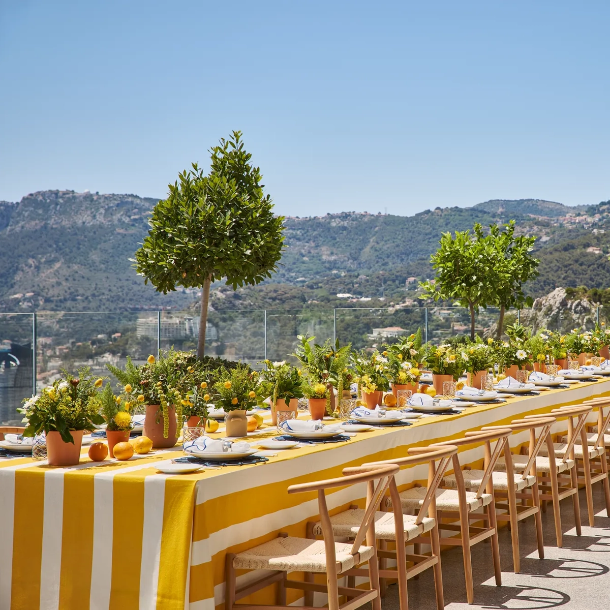 Long outdoor dining table set with yellow linens, wooden chairs, potted plants, and mountain views on a sunny terrace.
